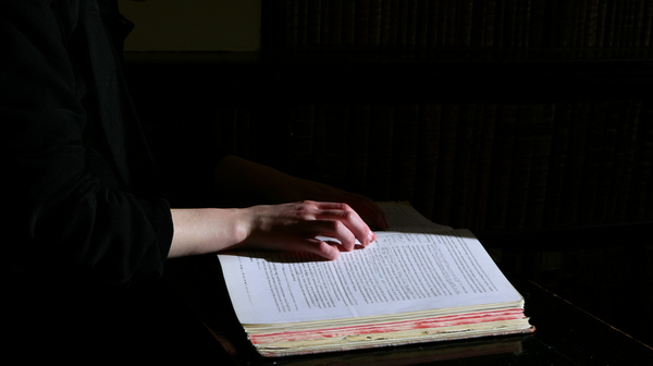 A dark and moody 16:9 cinematic horizontal shot inside an ancient London library with rich, dark wood tones. A detailed close-up of a character's…