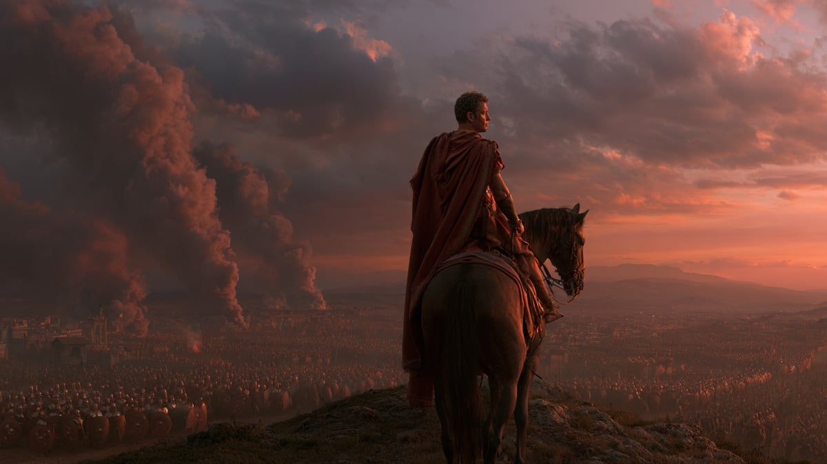 A monumental 16:9 vertical tracking shot across a vast ancient Roman battlefield at sunset. Thousands of legionaries with curved shields are arrayed in formation. A general in a crimson-lined cloak rides a powerful horse up a hill, looking down at the dramatic conflict and columns of smoke. High-contrast amber light, detailed textures, sweeping scale, photorealistic masterpiece